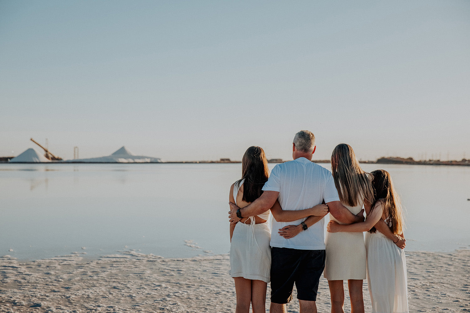 A family standing arm in arm on the shore of a still bay at dusk, looking out over the water.