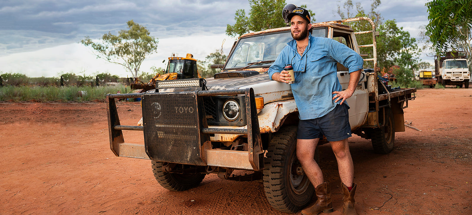 A station worker leaning against a dusty LandCruiser ute in red outback dirt, holding a Fridgy bottle.