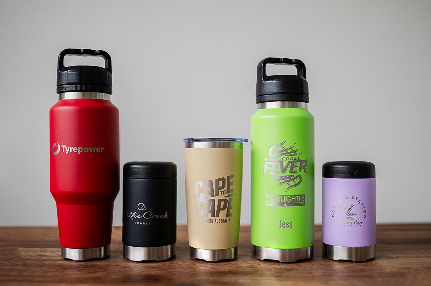 Five co-branded Fridgy bottles and can coolers lined up on a bench, each laser engraved with a different company logo.