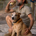 Man in a hat and khaki shirt holding a 600ml tumbler sitting with a dog, both on a dirt path.