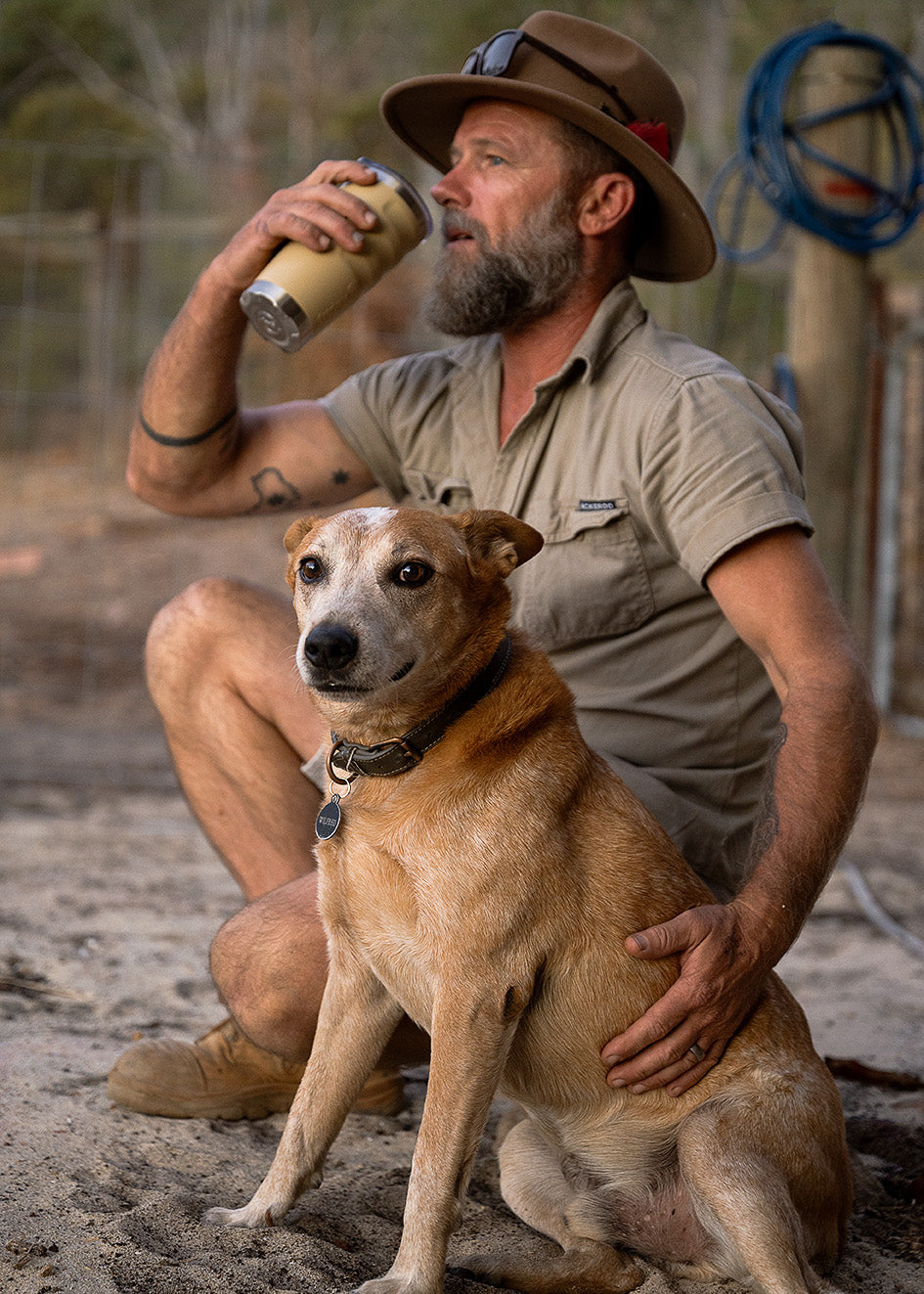 Man in a hat and khaki shirt holding a 600ml tumbler sitting with a dog, both on a dirt path.