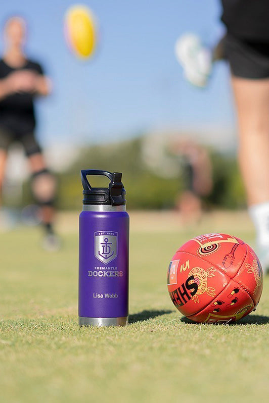 Fridgy 1080ml Engraved Grip Range Bottle next to a footy at the Fremantle Dockers women's training session – Double-wall vacuum insulated stainless steel water bottle built for durability and all-day hydration.