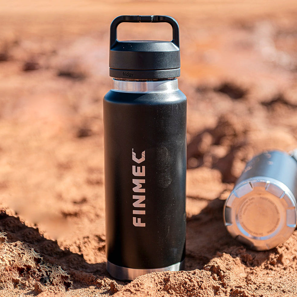 Fridgy 780ml Grip Range Bottle in Dark Hour Black with custom engraved logo – One bottle standing upright in red dirt, another resting on its side. Triple-wall insulated stainless steel water bottles built for durability and all-day hydration in rugged conditions.