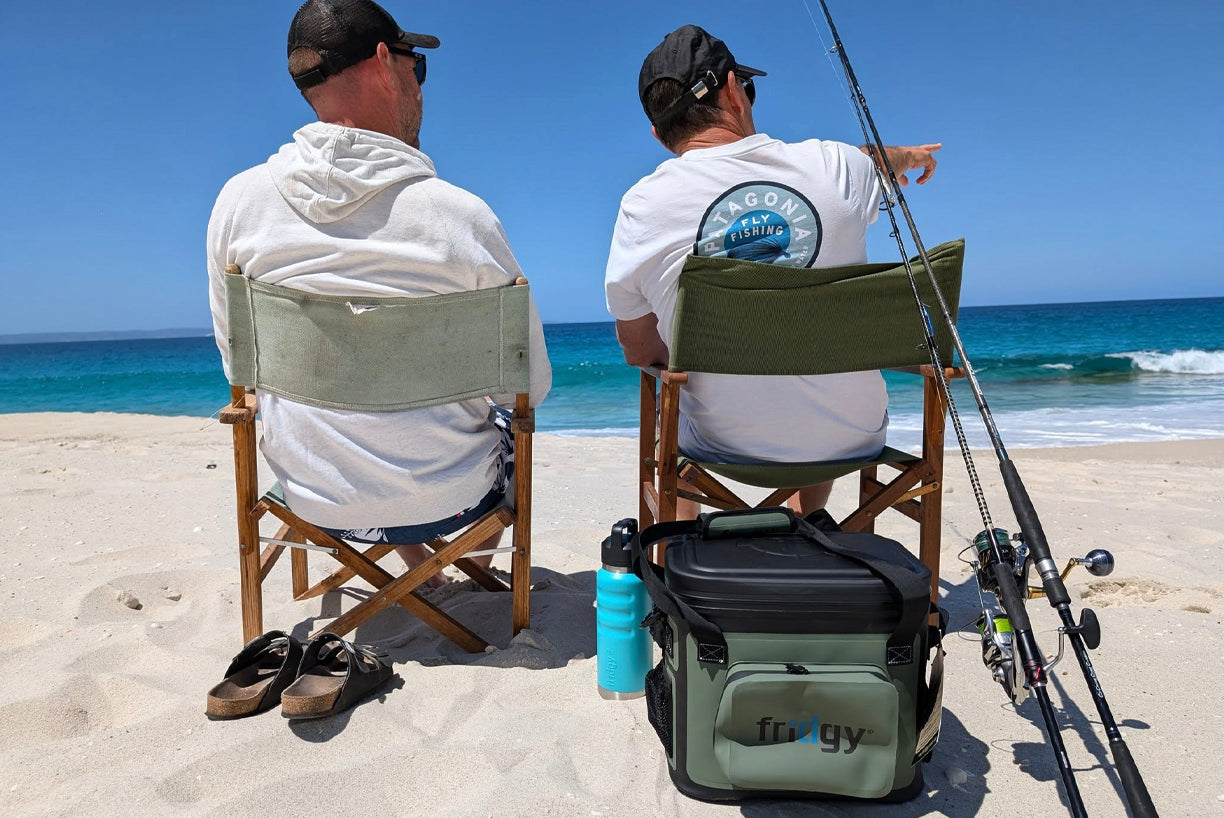 Fridgy CribMate Soft Cooler in the sand behind two fishermen on the beach – Insulated lunch cooler designed to keep food and drinks fresh during long fishing trips.