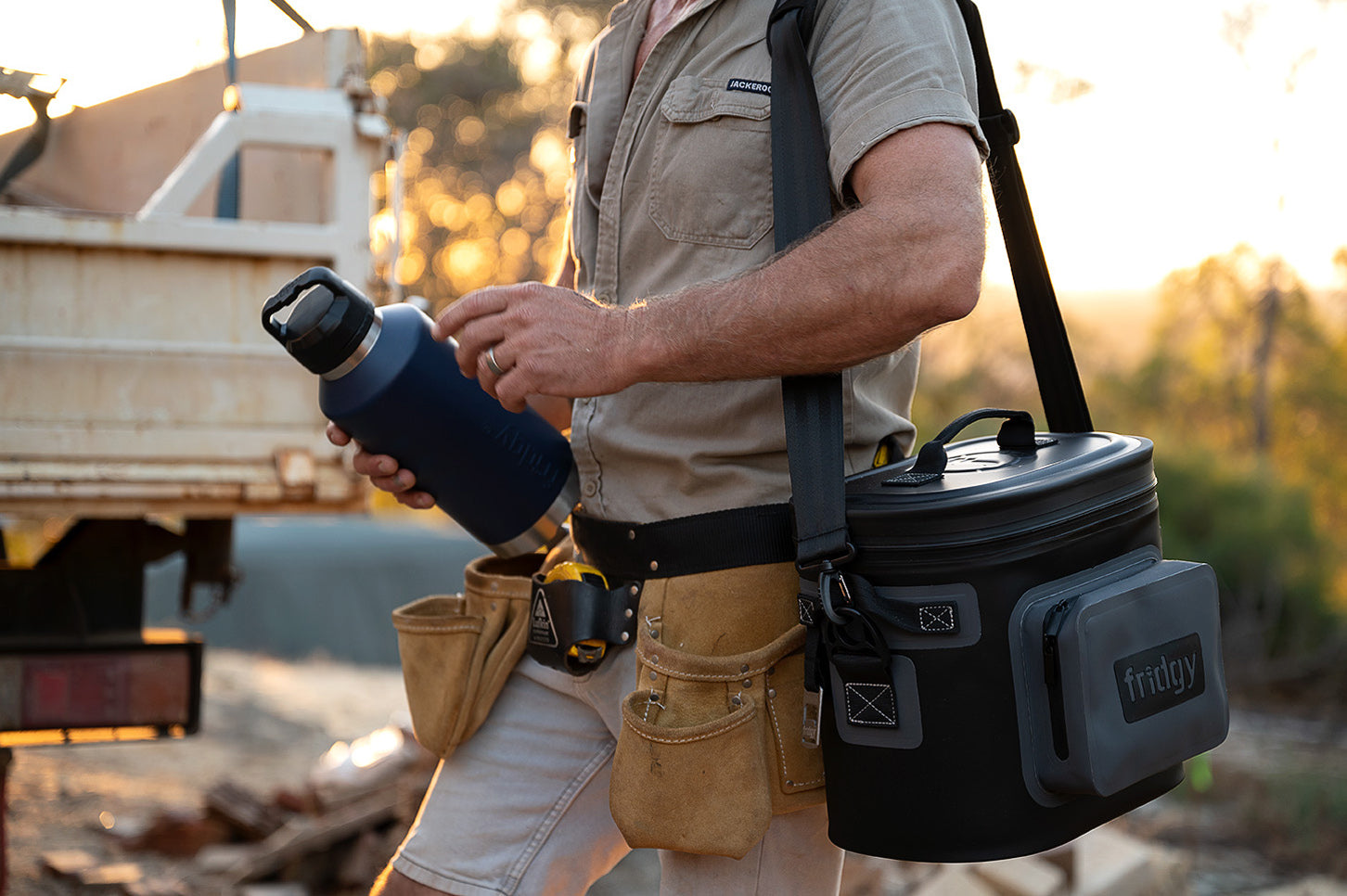 Man carrying Fridgy CribMate Cooler in Slate Grey over his shoulder and holding a 1.9L All-Dayer Bottle in Navy Blue – Built for rugged use and all-day hydration on the go.