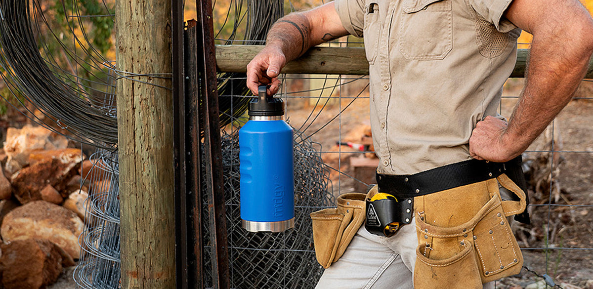 A tradie in workwear leaning on a fence, holding a blue Fridgy Grip Range insulated bottle, tool belt on.