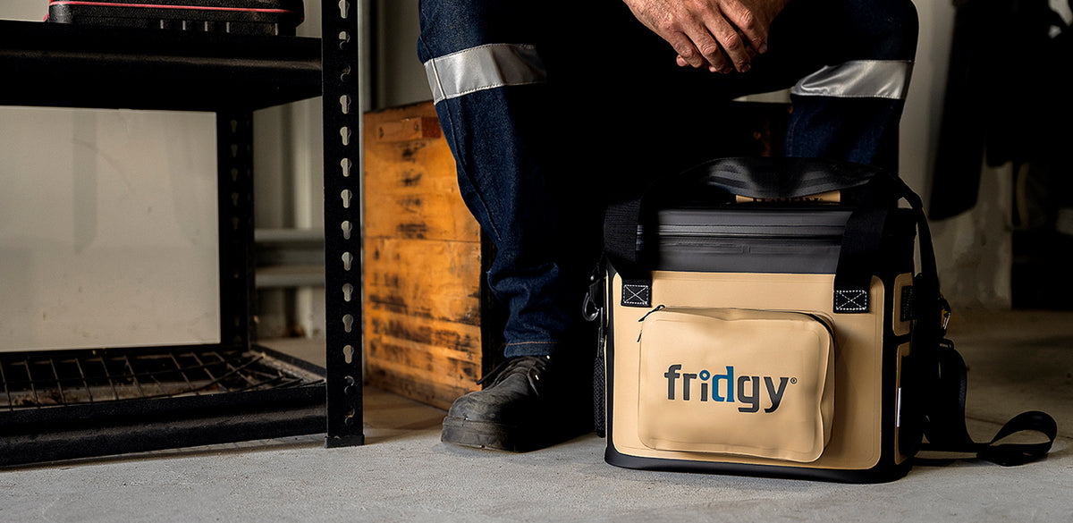 A tan and black Fridgy soft cooler bag on a shed floor beside a worker in hi-vis trousers and steel-cap boots.